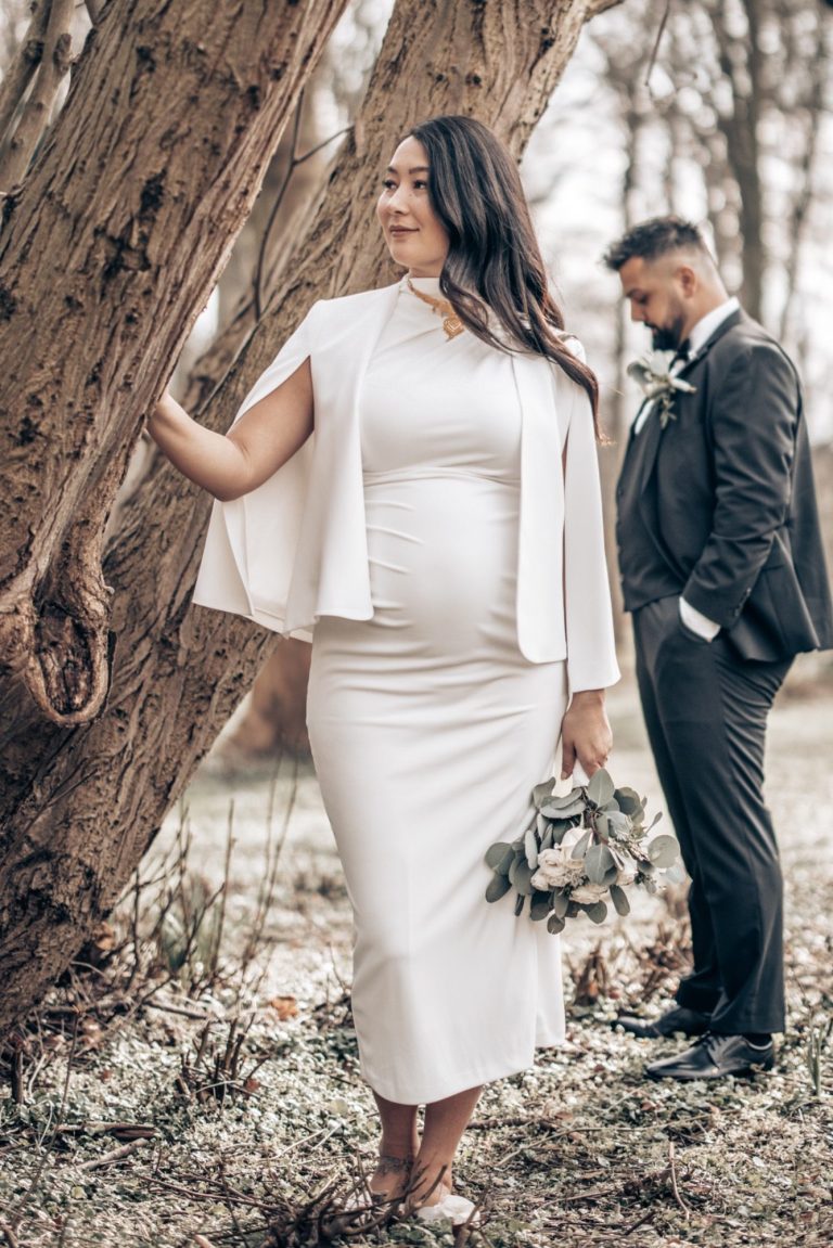 Schwangere Frau in elegantem Kleid mit Brautstrauß, im Hintergrund ein Mann im Anzug.