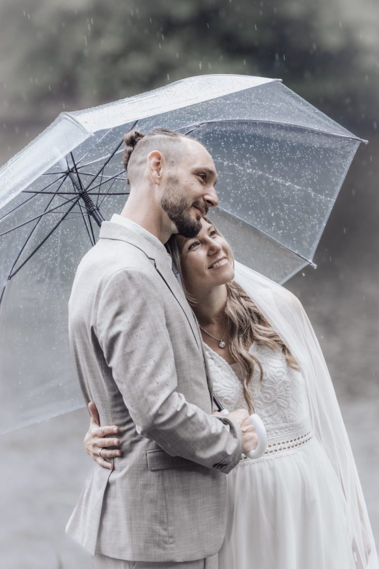 Ein Brautpaar steht unter einem Regenschirm und umarmt sich romantisch im Regen.